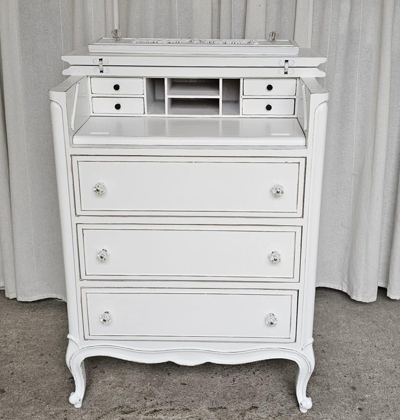 May include: A white, vintage-style secretary desk with a drop-down writing surface. The desk features three drawers with crystal knobs, small interior drawers, and a storage compartment. The desk has a curved base and ornate detailing, and is painted white with silver trim.