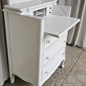 May include: White antique desk with a flip-up top and pull-out writing surface. The desk has three drawers with crystal knobs and decorative curved legs. The interior features small compartments and drawers for storage.