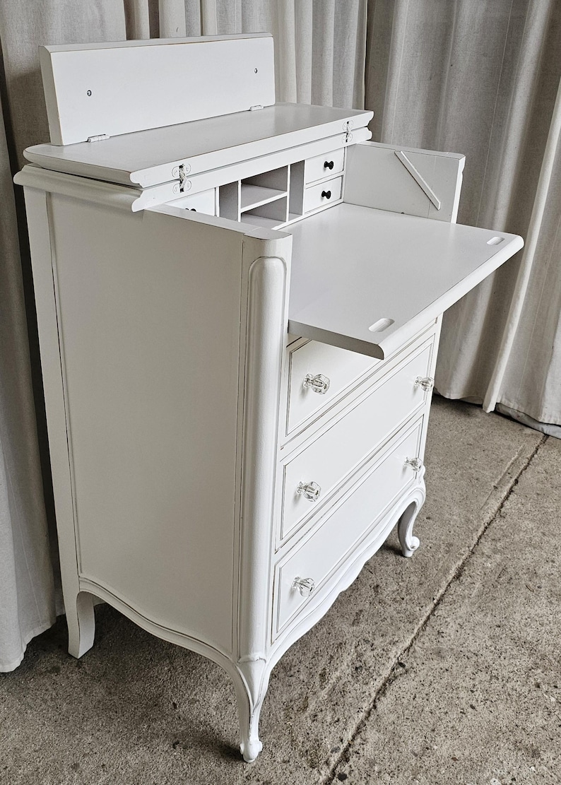 May include: White antique desk with a flip-up top and pull-out writing surface. The desk has three drawers with crystal knobs and decorative curved legs. The interior features small compartments and drawers for storage.