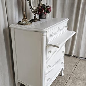 May include: A white antique dresser with three drawers and a pull-out shelf. The dresser has crystal knobs and decorative trim. A vintage mirror, a small tray, and a vase of flowers sit on top. The dresser is a classic piece of furniture.