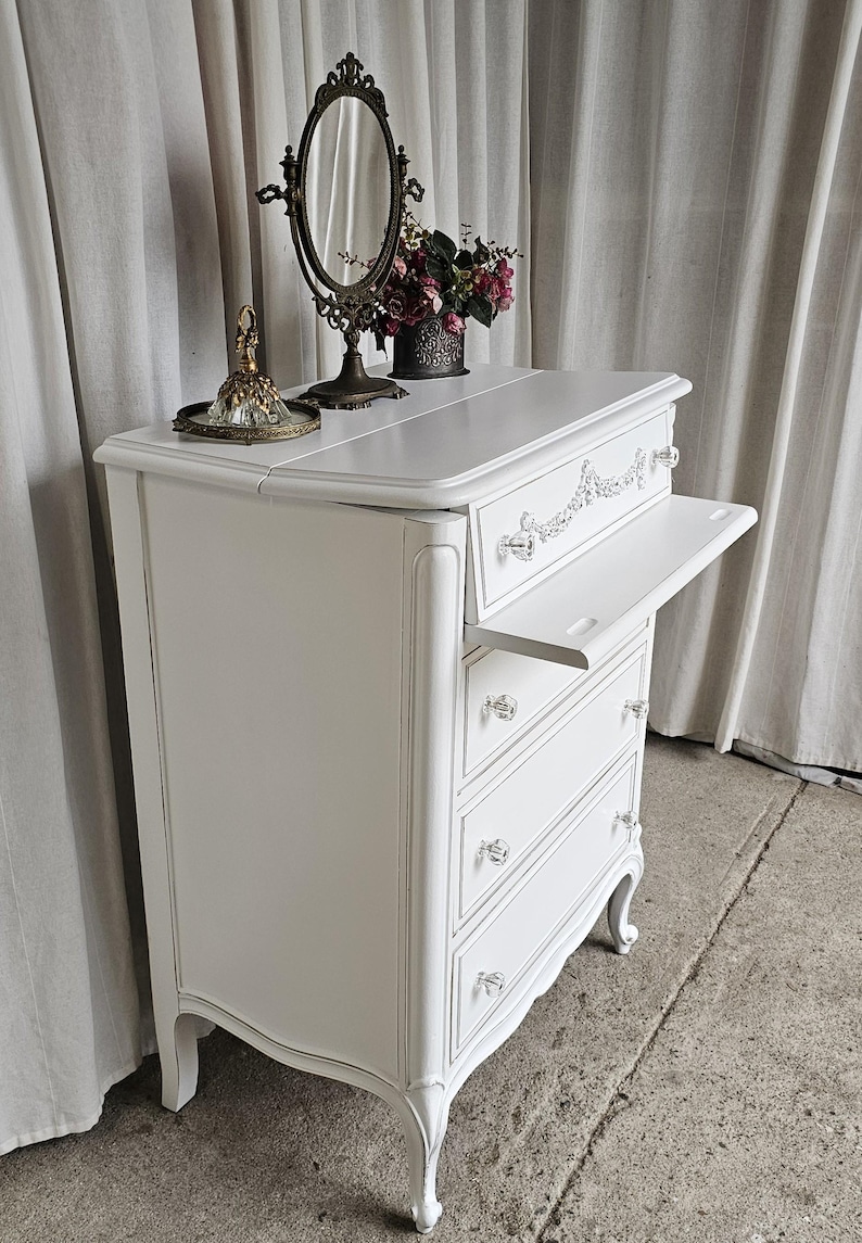 May include: A white antique dresser with three drawers and a pull-out shelf. The dresser has crystal knobs and decorative trim. A vintage mirror, a small tray, and a vase of flowers sit on top. The dresser is a classic piece of furniture.