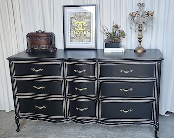 May include: Black dresser with nine drawers, gold hardware, and a decorative gold trim. The dresser has a curved front and sits on curved legs. A framed artwork, a small wooden box, a plant, and a decorative lamp are on top.