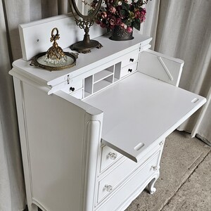 May include: White antique vanity desk with a fold-down writing surface, drawers, and crystal knobs. The desk features a decorative mirror, a tray with a glass perfume bottle, and a vase of flowers. The desk is painted white.