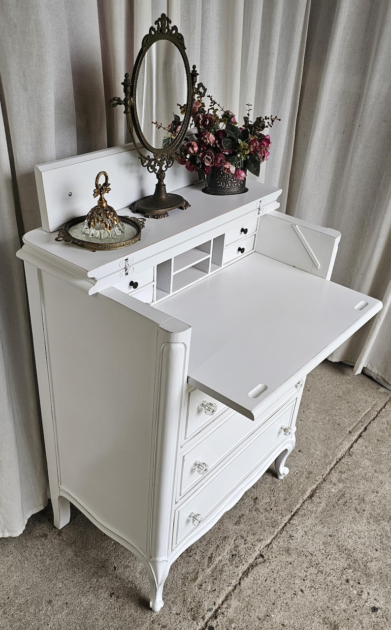 May include: White antique vanity desk with a fold-down writing surface, drawers, and crystal knobs. The desk features a decorative mirror, a tray with a glass perfume bottle, and a vase of flowers. The desk is painted white.