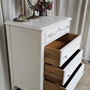 May include: White vintage dresser with three open drawers. The dresser has crystal knobs and decorative trim. A vanity mirror, perfume bottle, and flowers sit on top. The drawers are made of wood.