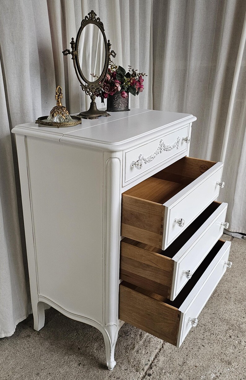May include: White vintage dresser with three open drawers. The dresser has crystal knobs and decorative trim. A vanity mirror, perfume bottle, and flowers sit on top. The drawers are made of wood.