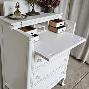 May include: A white vintage vanity desk with a fold-down writing surface and three drawers. The desk features crystal knobs, ornate legs, and a decorative mirror. A small tray with a perfume bottle and a vase of flowers are on top.