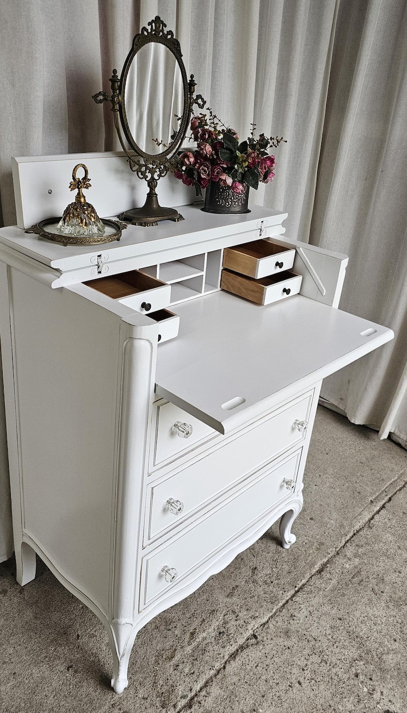 May include: A white vintage vanity desk with a fold-down writing surface and three drawers. The desk features crystal knobs, ornate legs, and a decorative mirror. A small tray with a perfume bottle and a vase of flowers are on top.