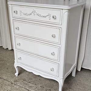May include: A white painted chest of drawers with four drawers and crystal knobs. The top drawer features a decorative floral design. The chest has curved legs and a distressed finish, giving it a vintage look.