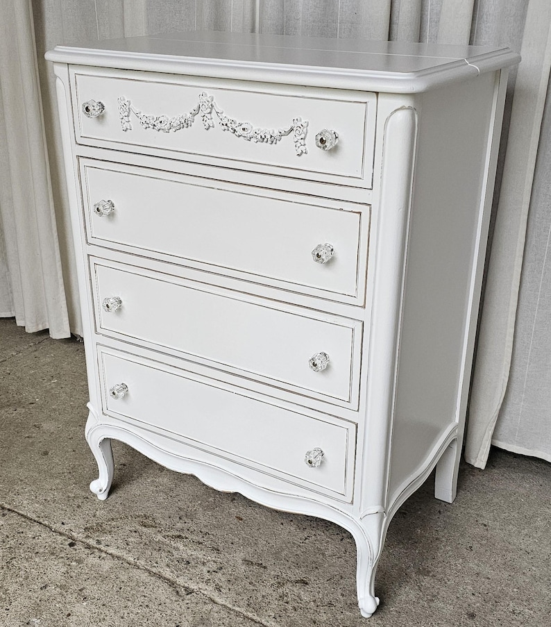 May include: A white painted chest of drawers with four drawers and crystal knobs. The top drawer features a decorative floral design. The chest has curved legs and a distressed finish, giving it a vintage look.