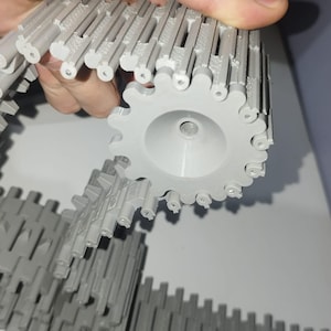 Op de afbeelding: Een lichtgrijze 3D-geprinte tankketting- en wielconstructie. Het wiel heeft een centraal gat en een getande buitenrand. De ketting bestaat uit in elkaar grijpende rechthoekige segmenten. De onderdelen worden door een hand vastgehouden.