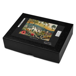 May include: A black box with a colourful jigsaw puzzle image on the front. The puzzle depicts a white cottage with a red roof surrounded by a lush garden with flowers in shades of red, pink, yellow, and orange. The text on the box reads "Puzzle 1000 520 Pcs. LOT 231968160 Made in USA."