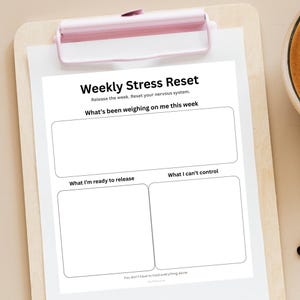 Może przedstawiać: Biała kartka z tekstem "Weekly Stress Reset" i zachętami do pisania o tygodniowych czynnikach stresogennych. Papier jest trzymany przez różowy schowek na drewnianej powierzchni. Zachęty obejmują "What's been weighing on me this week".