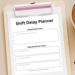 Może przedstawiać: Biały Shift Delay Planner na drewnianej podkładce z różowym klipsem. Planer zawiera sekcje Delayed Start Focus i Adjusted Plan for Today, z monitami do refleksji i planowania.