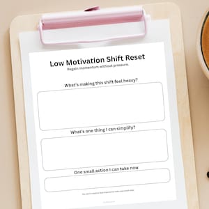 Może przedstawiać: Biała kartka z tekstem "Low Motivation Shift Reset" i pytaniami do autorefleksji, trzymana przez różową tablicę. Pytania obejmują kwestie dotyczące tego, co sprawia, że zmiana wydaje się ciężka i jakie działania należy podjąć. Tablica spoczywa na drewnianej powierzchni.