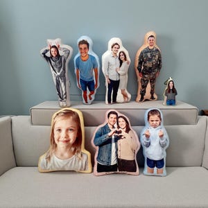 May include: A collection of custom-printed body pillows featuring various people. The pillows display full-body images of individuals in different outfits and poses. The pillows are arranged on a gray sofa, showcasing a variety of personalized designs.