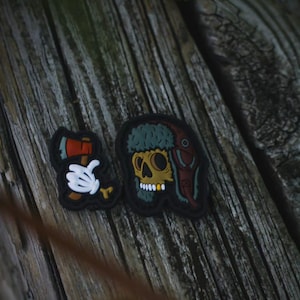 Puede incluir: Dos parches sobre una superficie de madera. Un parche presenta un hacha de dibujos animados con una mano enguantada blanca. El otro parche es una calavera con un sombrero de piel con un diseño rojo y marrón.
