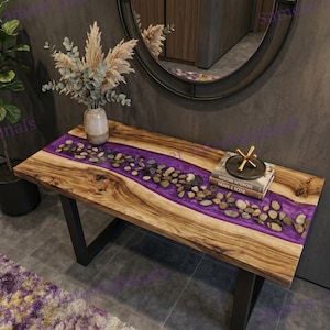 May include: A rectangular console table featuring a natural wood grain surface with a central river-like inlay of translucent purple resin embedded with smooth river stones. The table is supported by black metal legs and holds a vase with dried foliage.