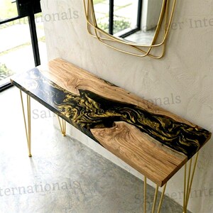 May include: A rectangular console table featuring a natural wood slab top with a central river-like inlay of black and gold swirling resin. The table is supported by four thin, gold-colored metal hairpin legs and stands against a textured wall.