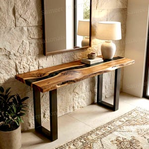 May include: A rectangular console table featuring a natural live-edge wood slab with a dark resin river inlay, supported by two black metal U-shaped legs. A white table lamp and a small bowl sit on the surface of the wooden console table.