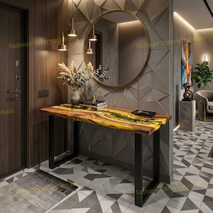 May include: A rectangular console table featuring a natural wood slab top with a central yellow and black resin river inlay, supported by two black metal U-shaped legs. The table is positioned against a wall with geometric gray panels.