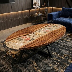 Könnte beinhalten: Ein ovaler Couchtisch mit zwei polierten Holzplatten und einer zentralen Harzeinlage, die mit Sand, verschiedenen Muscheln und Seesternen gefüllt ist. Der Tisch wird von einem schwarzen, X-förmigen Metallgestell getragen.