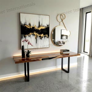 May include: A rectangular console table with a polished wood grain top and black metal legs, featuring a square abstract painting in black, white, and gold, a round gold-framed mirror, a black vase with pink flowers, and a stack of books.