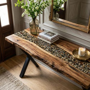 May include: A live edge wooden console table with a central inlay of smooth river stones encased in clear resin. The table features black metal X-shaped legs and holds a glass vase with white flowers, three stacked books, and a candle on a gold tray.