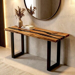 May include: A rectangular console table featuring a natural wood slab top with a dark, flowing resin inlay running through the center. The table is supported by two black metal U-shaped legs and holds a ceramic vase with dried foliage and two books.
