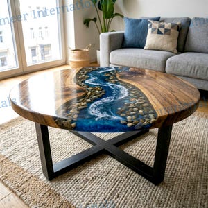 May include: Round coffee table with a unique river design. The table features a wooden surface with a blue epoxy resin river filled with small stones. It has a black metal base and sits on a beige rug.
