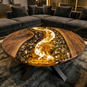 Op de afbeelding: Ronde salontafel met een uniek ontwerp. De tafel heeft een houten oppervlak met een rivier van hars en stenen in het midden. Hij heeft een zwart metalen onderstel en staat in een woonkamer.