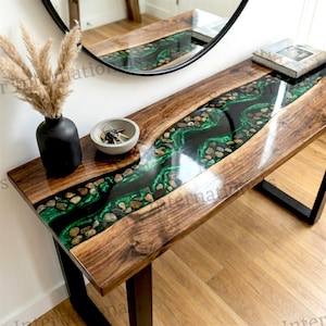 May include: A wooden console table with a river-like design. The table features a dark epoxy resin river filled with green stones, set within a walnut wood surface. The table has black metal legs and a glossy finish. A black vase and a bowl with keys are on the table.