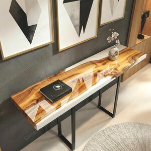 Puede incluir: Una consola moderna con una tapa de madera de borde vivo e incrustaciones de resina blanca. La mesa tiene una base de metal negro y un libro encima. La mesa mide aproximadamente 152 cm de largo.