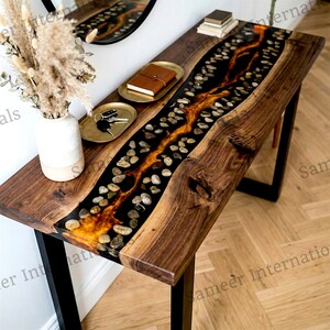 May include: A wooden console table with a river of black epoxy resin and embedded stones. The resin has a golden-orange glow. The table has a dark wood finish and black metal legs. A vase of dried flowers and a mirror are in the background.