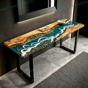 May include: A handcrafted wooden console table with a river-like design. The table features a live edge wood top with a turquoise epoxy resin river, filled with stones. The table is supported by black metal legs. A small tray and bowl are on the table.