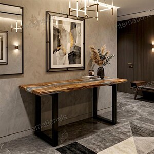 May include: A rectangular console table featuring a natural wood slab top with a central gray resin inlay, supported by two black metal U-shaped legs. The table is positioned against a wall with a framed abstract painting and a vase of dried grass.