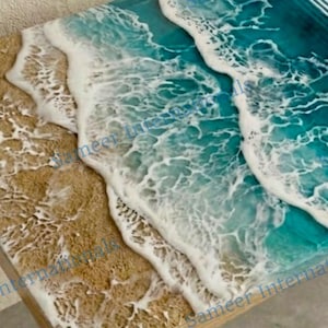 Può includere: Una superficie decorativa in legno con un design dettagliato di onde oceaniche. L'opera presenta un'area di spiaggia sabbiosa in tonalità beige e bianco, che sfuma in onde turchesi e bianche.