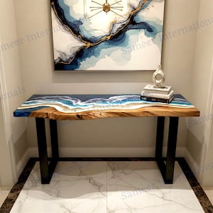 May include: A wooden console table with a live edge design. The tabletop features a blue and white resin design resembling ocean waves. The table has black metal legs and is placed against a wall with abstract art.