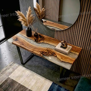 Puede incluir: Una consola de madera con un diseño de borde vivo y un río de resina epoxi transparente. La mesa tiene una base de metal negro y está decorada con un jarrón negro, plantas secas y una escultura dorada. La mesa mide aproximadamente 152 cm de largo.