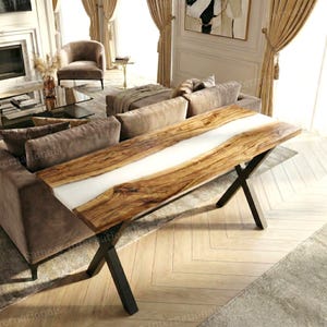 May include: A rectangular console table featuring a natural live-edge wood slab top with a central white resin inlay. The table is supported by a black metal X-shaped base, providing a stable foundation for the elongated wooden surface.