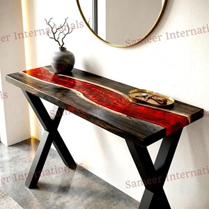 May include: A rectangular console table featuring a dark wood slab with a central, flowing river design made of vibrant red resin. The table is supported by black metal legs in an X-shape configuration. A small gold tray with keys sits on the surface.