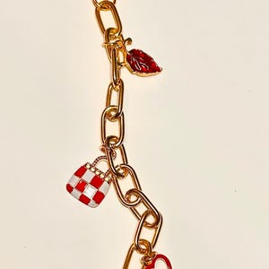 May include: Gold-toned chain with a lobster clasp, adorned with red and white enamel charms. Charms include a leaf, a picnic basket, a heart, and a strawberry. The chain is made of oval links.