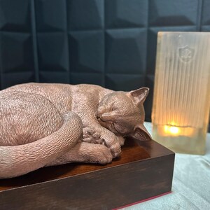 Könnte beinhalten: Eine bronzefarbene Katzenskulptur in Schlafposition, ruhend auf einem dunklen Holzsockel. Der Schwanz der Katze kringelt sich um ihren Körper. Eine brennende Kerze in einem Glashalter befindet sich im Hintergrund.