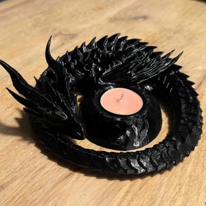 Pode incluir: Uma escultura de dragão preto enrolado, concebida como um suporte para velas. O dragão apresenta escamas detalhadas, chifres afiados e uma cauda curva que rodeia uma abertura circular central contendo uma pequena vela redonda cor de pêssego.