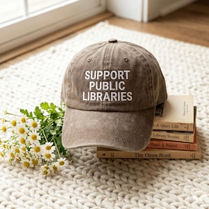 May include: A brown baseball cap with the white embroidered text SUPPORT PUBLIC LIBRARIES on the front panel. The cap features a structured crown, a curved brim, and a button on top, resting near a stack of books and a bunch of white daisies.