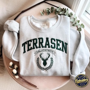 May include: A light grey crewneck sweatshirt featuring dark green embroidery. The text reads TERRASEN in an arched font above a banner with the word GALATHYNIUS. Below the banner is a dark green embroidered stag head framed by a laurel wreath.