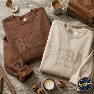 Op de afbeelding: Een bruine sweater en een crèmekleurige sweater, beide voorzien van geborduurde tekst BCBA EST. 2026 op de borst. De bruine mouw toont de naam Kelsey, de crèmekleurige mouw toont de naam Liam, beide met een klein hartvormig icoontje.