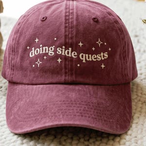 May include: A maroon baseball cap featuring the white embroidered text "doing side quests" across the front panel. The design includes small white star and dot accents surrounding the lettering, with a curved brim and stitched eyelets.