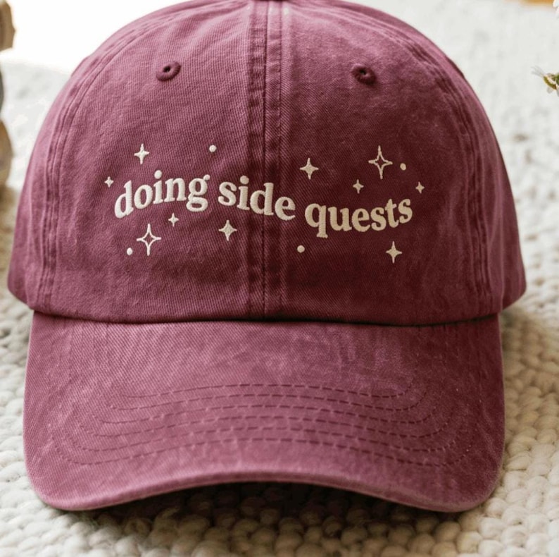 May include: A maroon baseball cap featuring the white embroidered text "doing side quests" across the front panel. The design includes small white star and dot accents surrounding the lettering, with a curved brim and stitched eyelets.