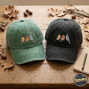 Op de afbeelding: Twee baseballpetten, één in saliegroen en één in antracietgrijs, elk voorzien van een borduursel van drie kleine vogels in geel, oranje en blauw. De petten hebben een verwassen textuur en een gestructureerde kroon met zes panelen.
